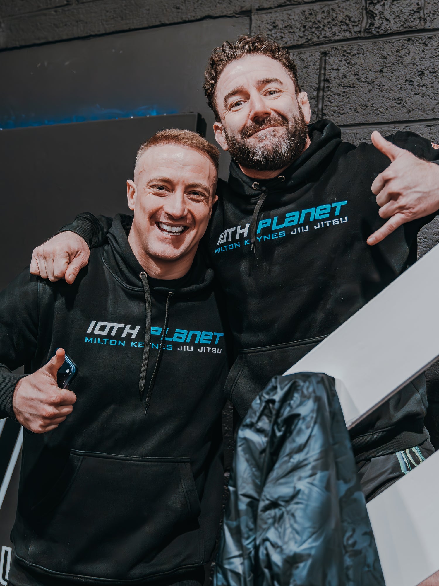 Two men wearing black hoodies with '10th Planet' branding posing in front of a black wall.