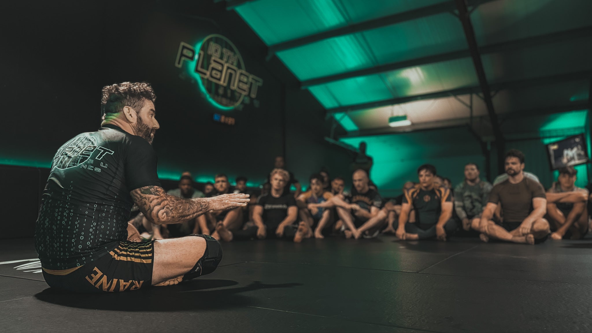 Head coach addressing a group of people in a bjj gym setting with '10th planet milton keynes' branding.