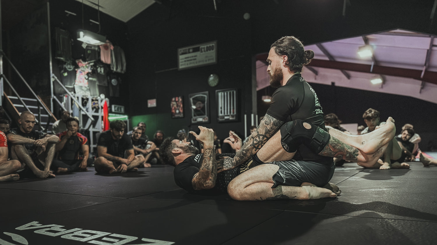 Two men practicing martial arts on a mat with spectators in the background.